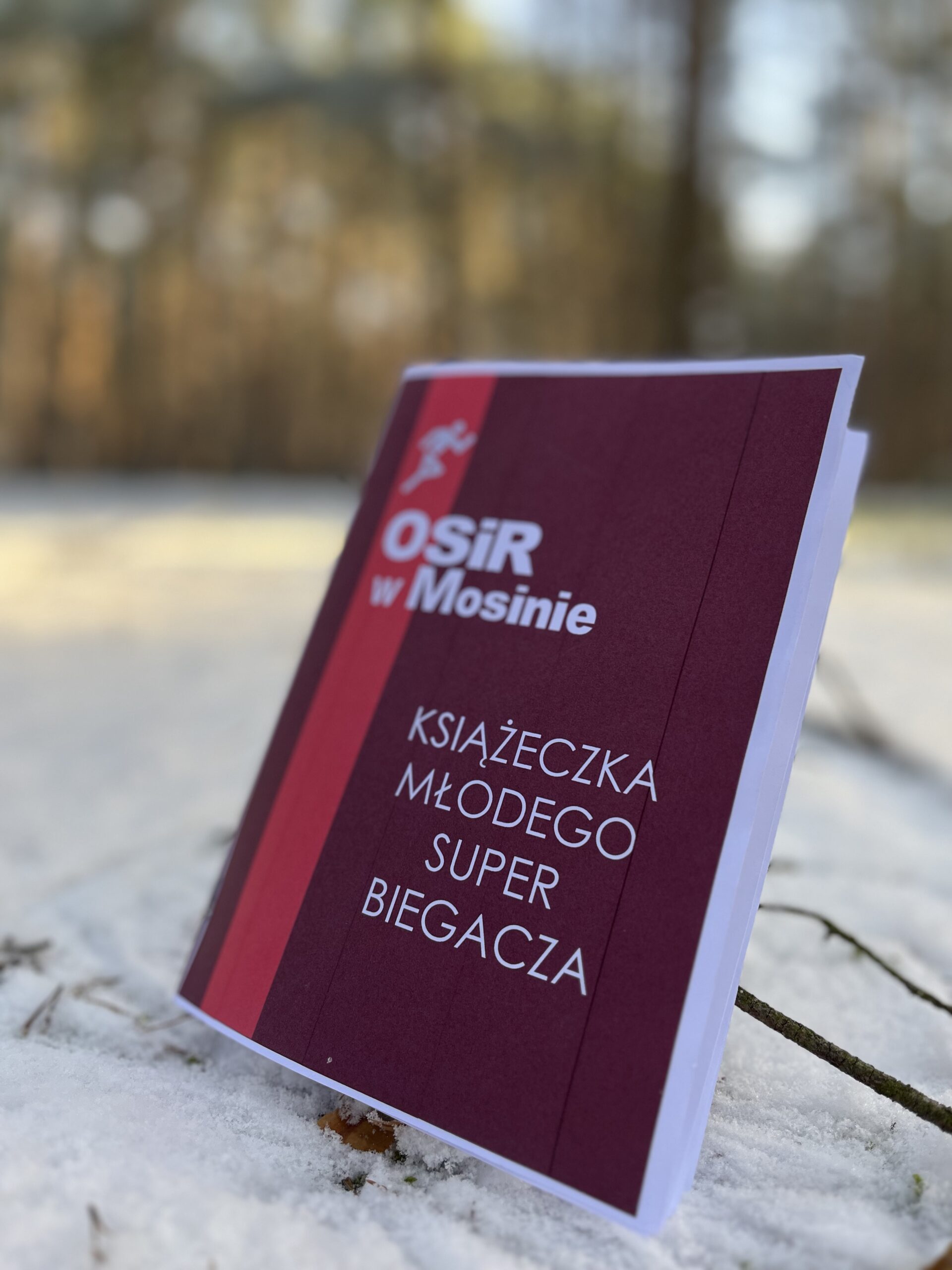 Książeczka Młodego Super Biegacza na tle zimowego lasu. Tekst w kolorze białym. Książeczka w kolorze bordowym. u góry, po lewej stronie logo ośrodka sportu i rekreacji w Mosinie